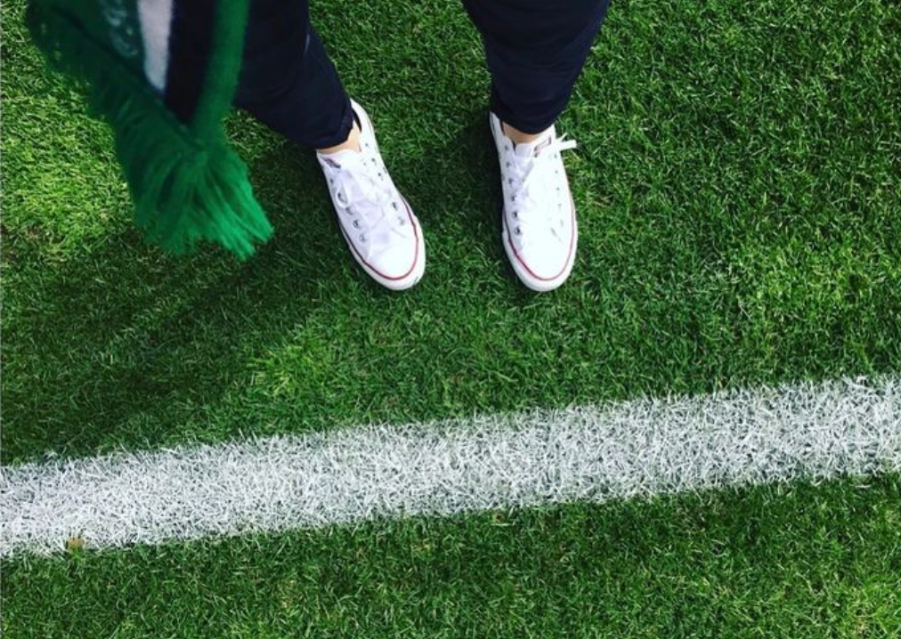 Close-up of white sneakers on vibrant artificial turf with a white line marking, suggesting a sports field or recreational area.